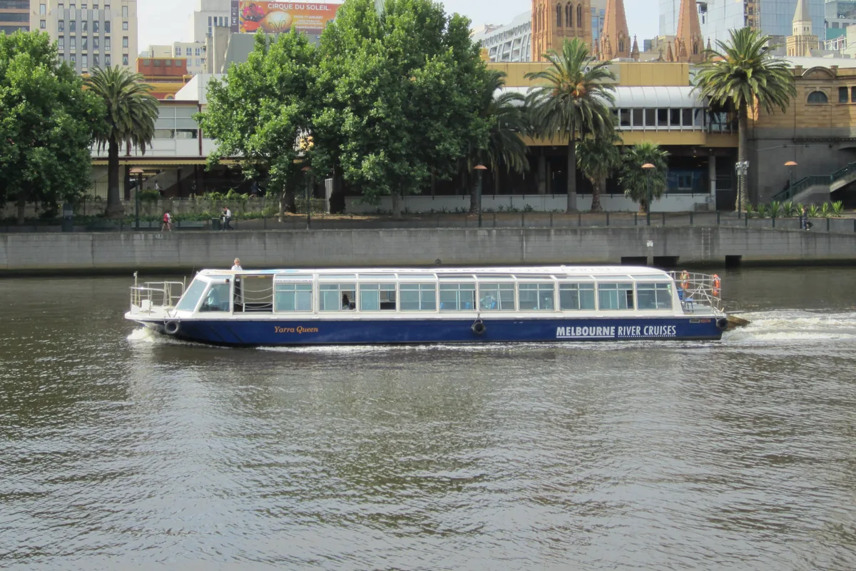 A white and blue river cruise boat on the Yarra beside trees and the roofs of the city's riverside buildings.