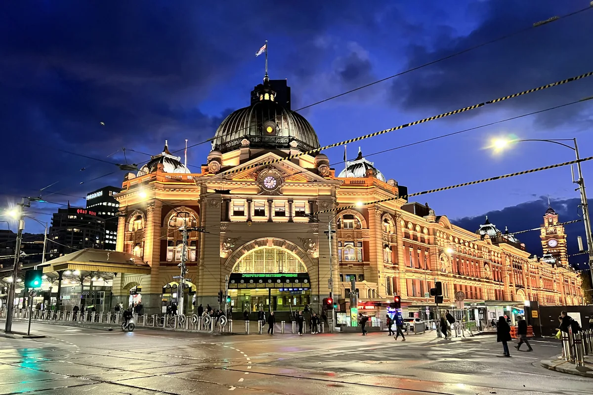 Flinders Street Station at night, its domed Edwardian building lit gold against a deep blue sky.